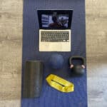 A computer on a video call, a foam roller, an exercise band, and a kettlebell. All on a yoga mat.