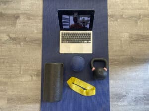 A computer on a video call, a foam roller, an exercise band, and a kettlebell. All on a yoga mat.