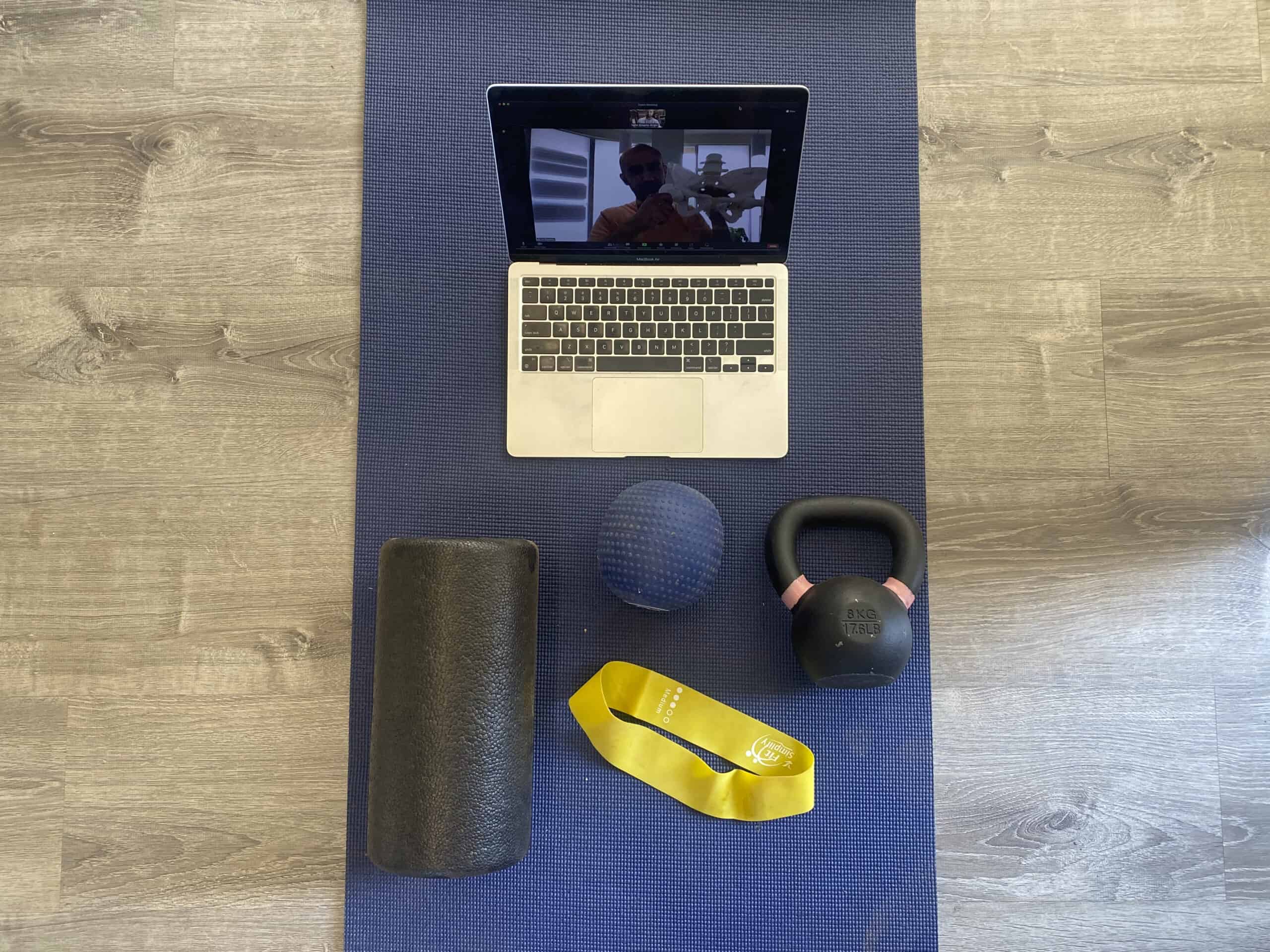 A computer on a video call, a foam roller, an exercise band, and a kettlebell. All on a yoga mat.